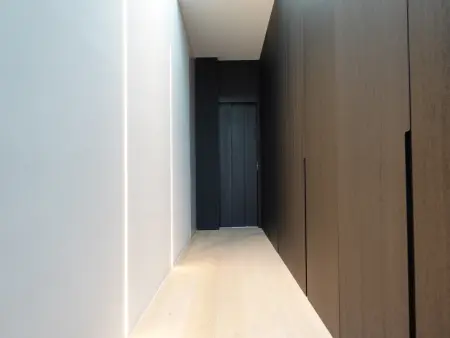 Hal met witte muren, geïntegreerde verlichting, lichthouten vloer en donkerhouten kasten met een donkere lift aan het einde.